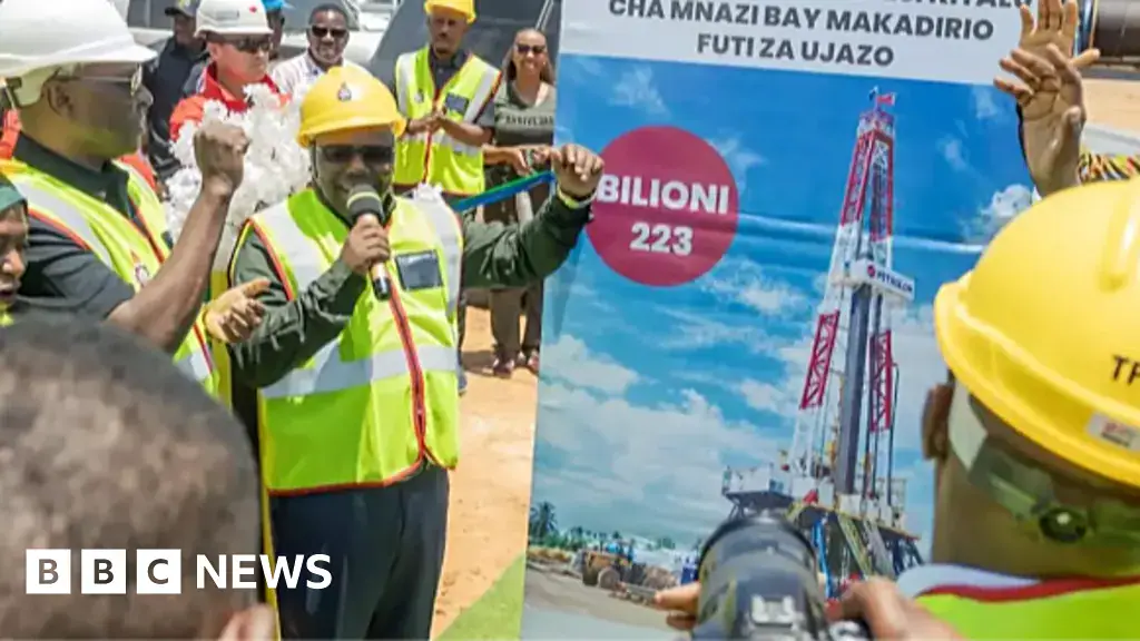 Energy Minister Juma Homera in yellow hard hat and yellow vest holds a microphone by a big poster as he announces to a small group and a photographer the discovery of 223 billion feet of natural gas.
