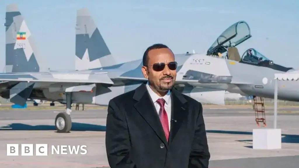 Abiy Ahmed, in a black suit and red tie standing on tarmac in front of a fighter jet
