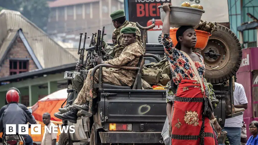 A woman wearing a red wrap skirt and a black shoulder bag walks away from a pick-up truck carrying M23 rebels in camouflage uniforms. She balances a grey basket of mangoes on her head.