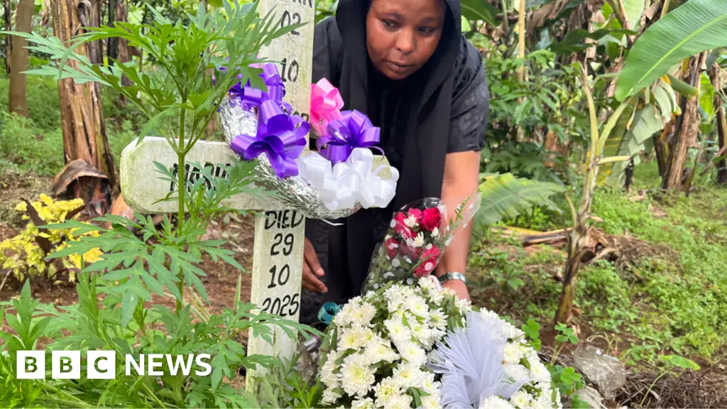 Neema Munuo attending a grave with a simple cross and the words 'Died 29 10 2025' written on it. There are flowers and ribbons on the cross in a densely vegetated area