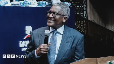 Aliko Dangote, wearing a suit, is smiling as he turns to the left of the picture. He is holding a microphone in his right hand.
