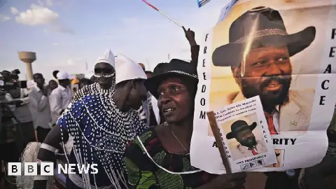 A crowd at a political rally holding a banner for South Sudan President Salva Kiir that reads "Freedom, peace and prosperity next to his portrait wearing a black cowboy hat