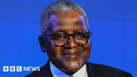 A man with grey hair, wearing a black suit, white shirt, and red tie, smiling into the camera.