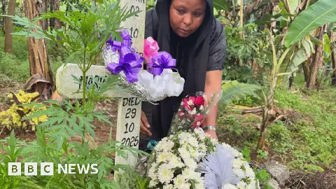 Neema Munuo attending a grave with a simple cross and the words 'Died 29 10 2025' written on it. There are flowers and ribbons on the cross in a densely vegetated area
