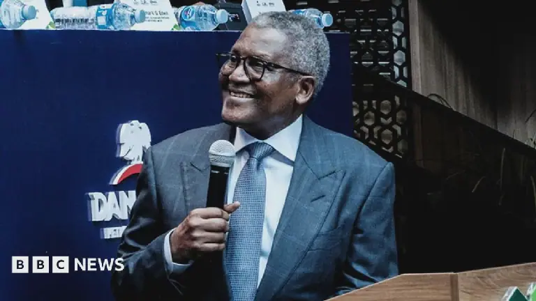 Aliko Dangote, wearing a suit, is smiling as he turns to the left of the picture. He is holding a microphone in his right hand.